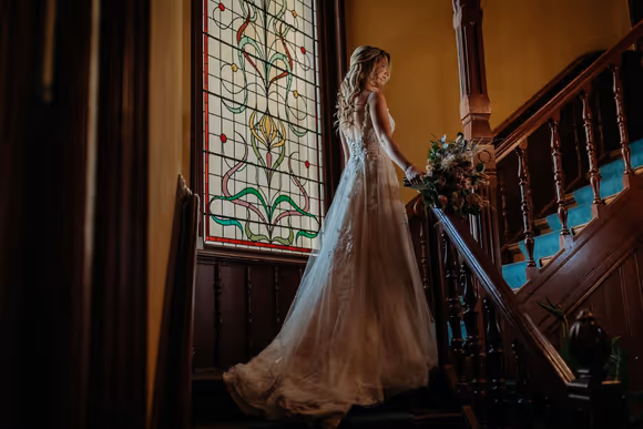 Bruid gaat trap op met een glas in lood raam op de achtergrond